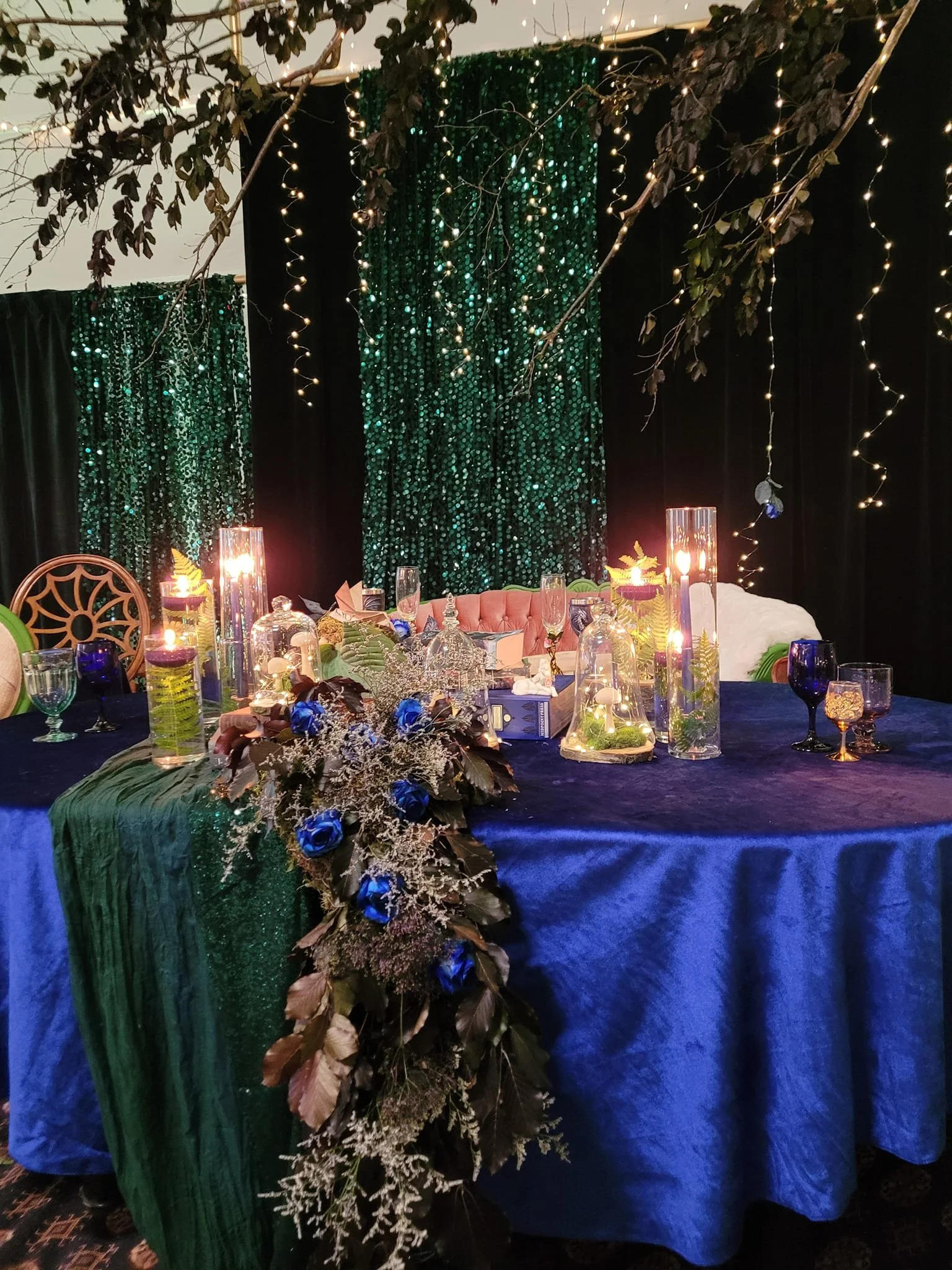 A decorated event table with a blue tablecloth, floral centerpiece with blue roses and greenery, tall glass vases with candles and flowers, and a backdrop of green sequin curtains and string lights, with hanging branches and leaves overhead.