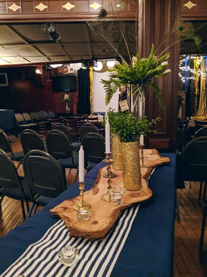 A table decorated with a natural wood centerpiece, gold candles in candlesticks, gold vases, and greenery in a gold glitter vase, set for a celebration or event in a banquet hall.