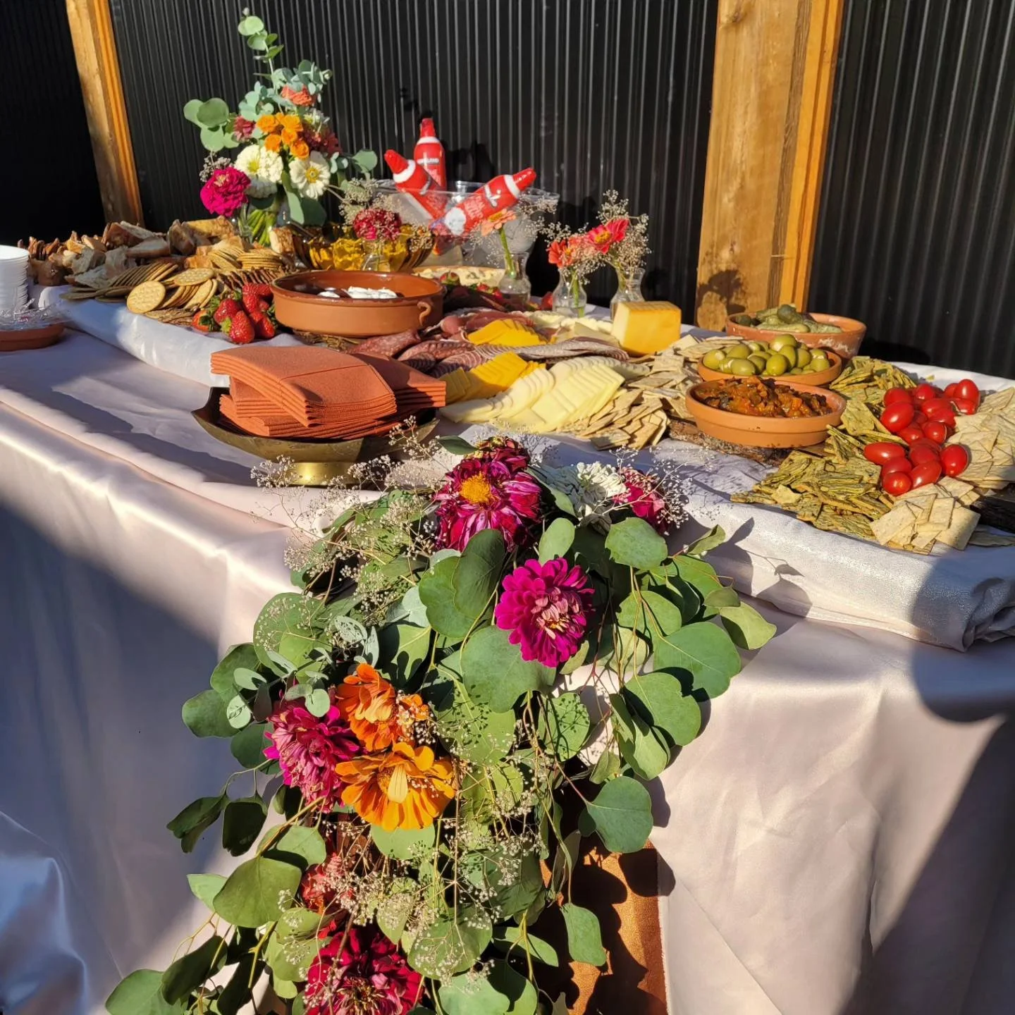 A long table set with various foods like cheese, olives, tomatoes, crackers, and meats, decorated with colorful flowers and floral arrangements for an outdoor buffet or celebration.