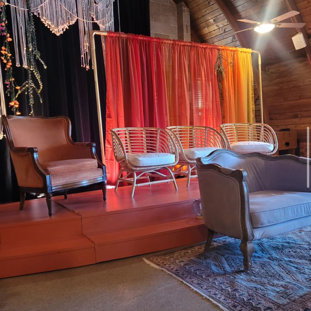 Indoor stage setup with vintage and wicker furniture and colorful curtains in shades of red, orange, and yellow, with a wooden wall and ceiling fan in the background.