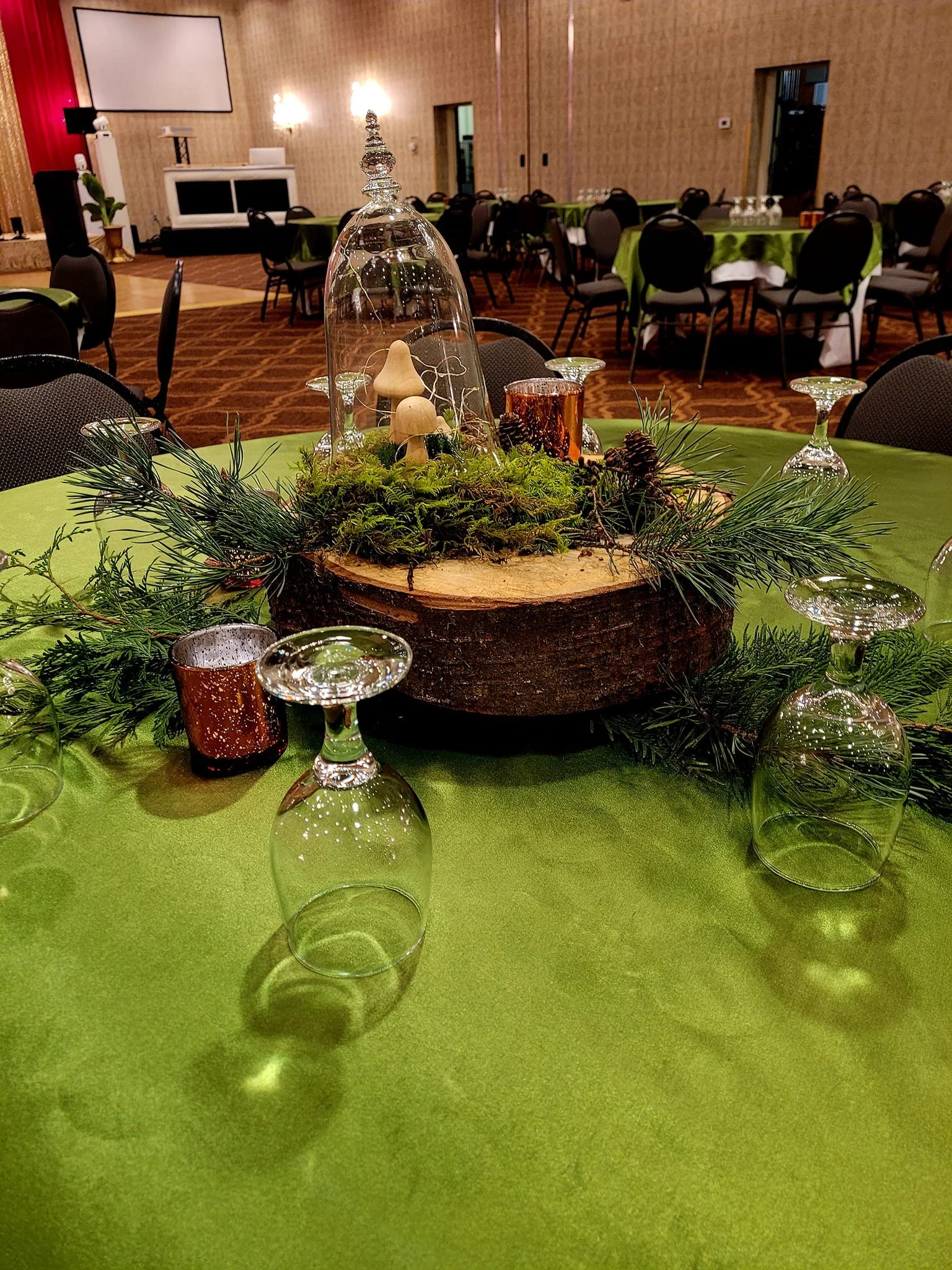 Table centerpiece with greenery, pinecones, a glass cloche with small mushrooms, and wooden decor on a green tablecloth in a decorated banquet hall.