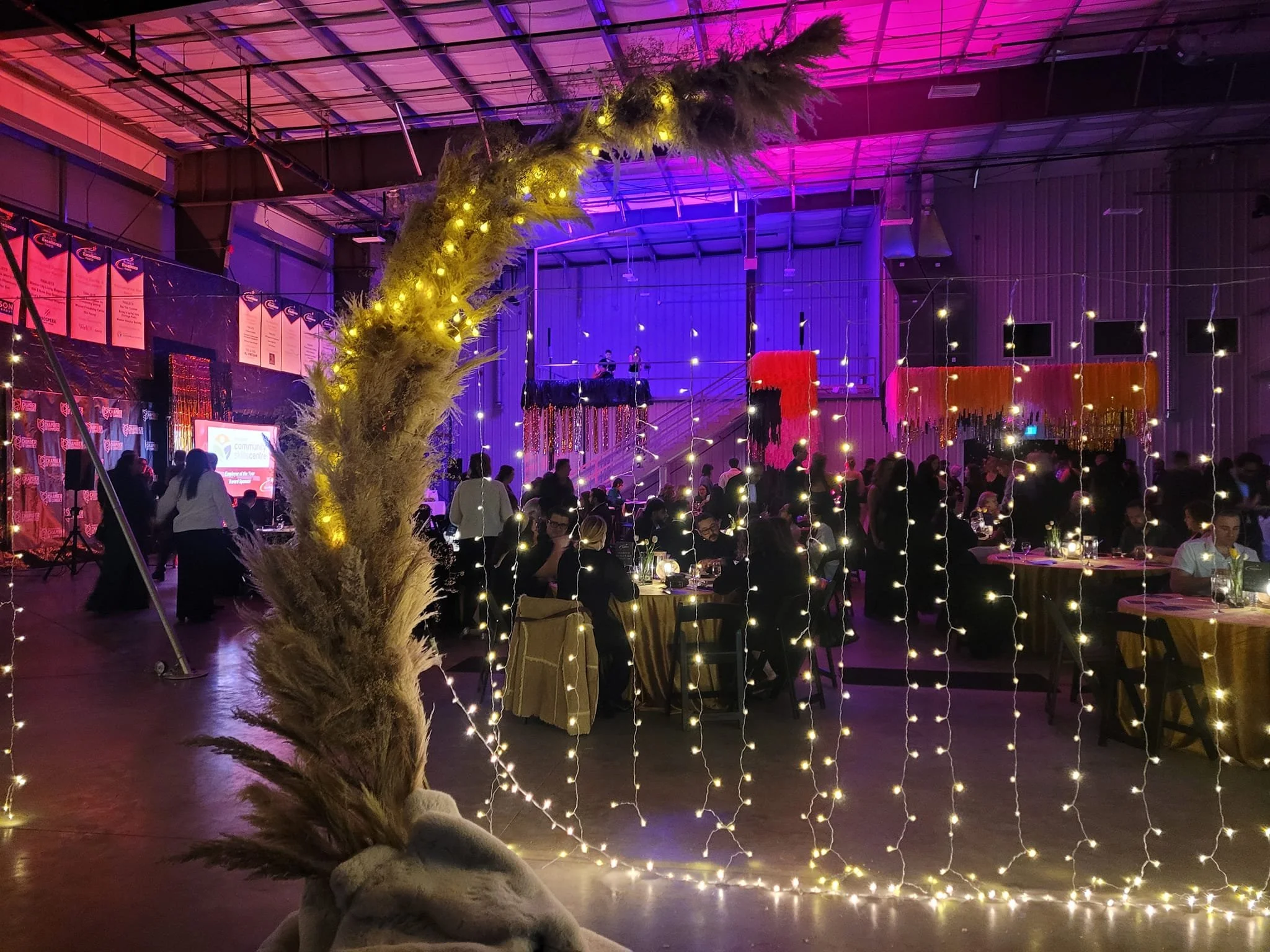 Indoor event with people dining at tables decorated with lights. Decorative hanging ribbons and a large illuminated arch in the foreground, with a stage and banners visible in the background.