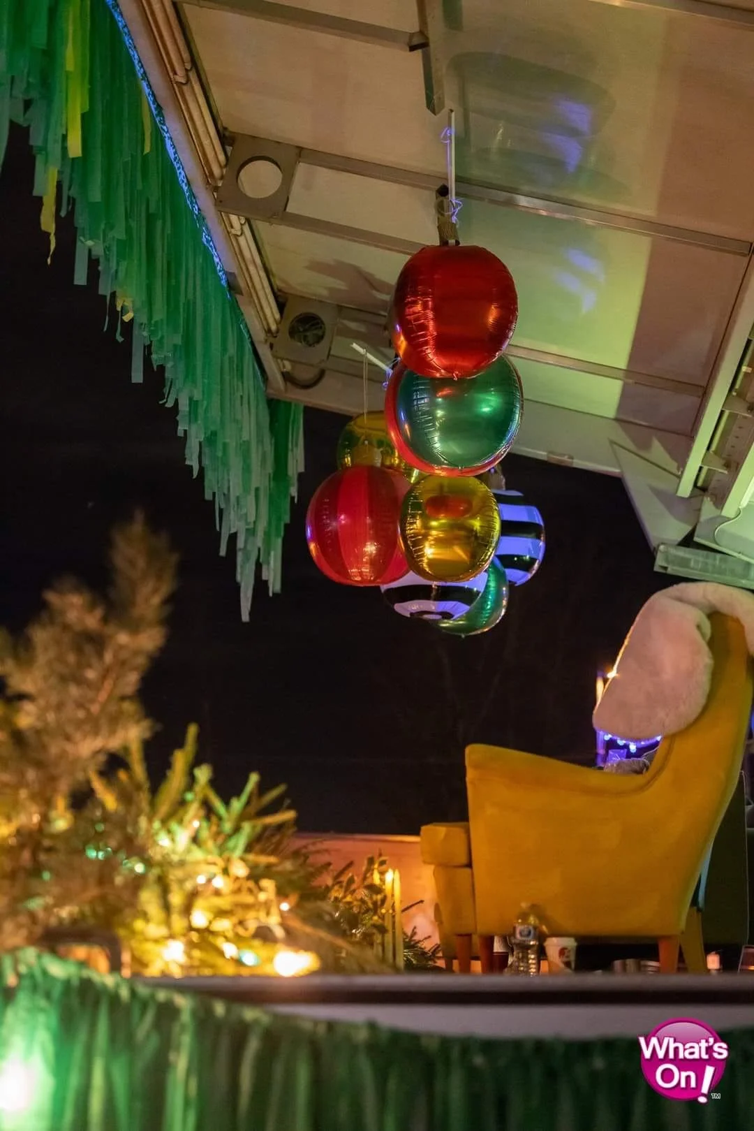 Colorful hanging paper lanterns in red, blue, green, and yellow, decorated for a festive event at night, with a yellow chair and some greenery in the foreground.