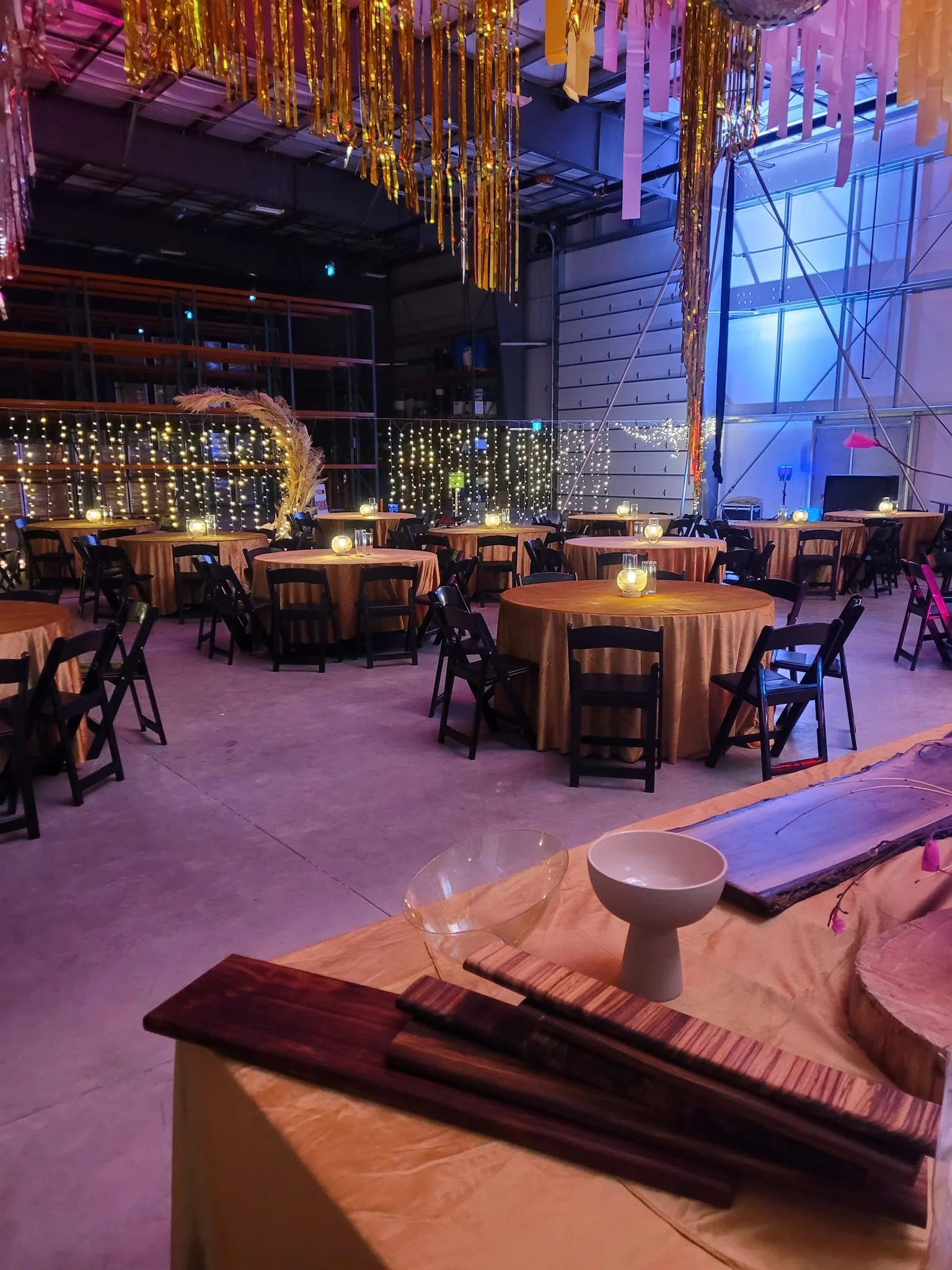 Event setup inside a large industrial space with round tables covered in tan tablecloths, black chairs, and decorative string lights and hanging gold and pink streamers.