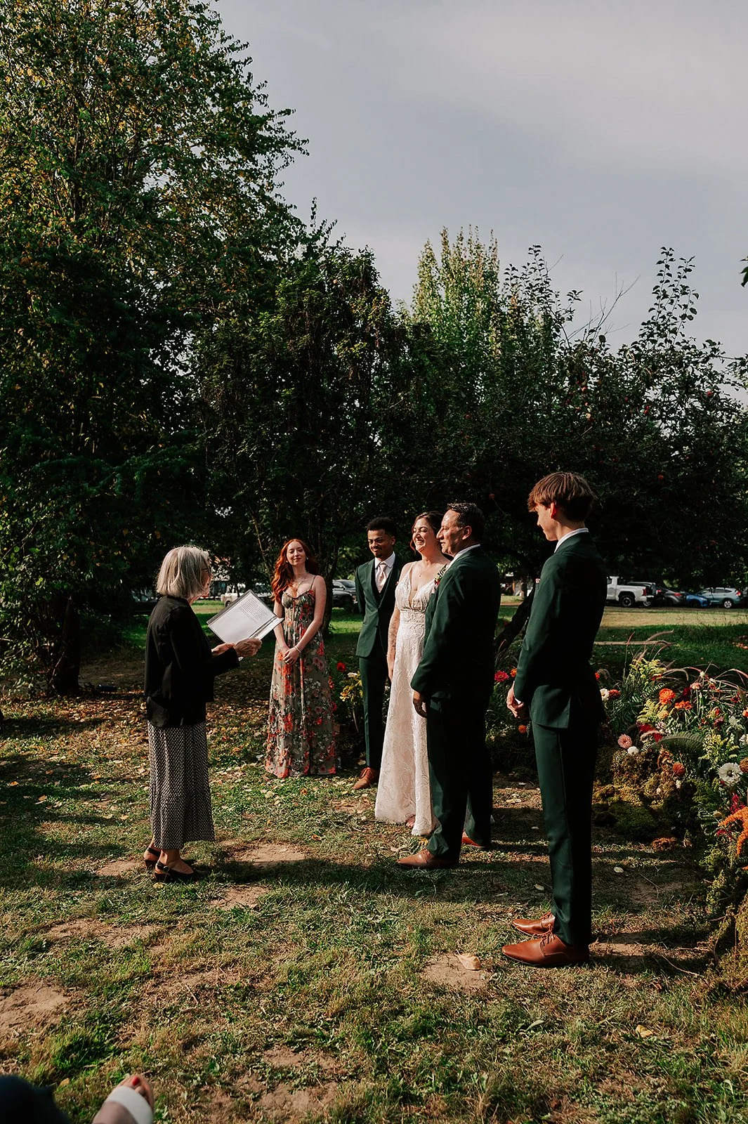 A wedding ceremony outdoors with five people standing in front of a large tree, with a woman officiating holding a book or paper, and the bride and groom along with two other attendants. The bride is wearing a white lace dress, and the groom and attendants are dressed in dark suits.