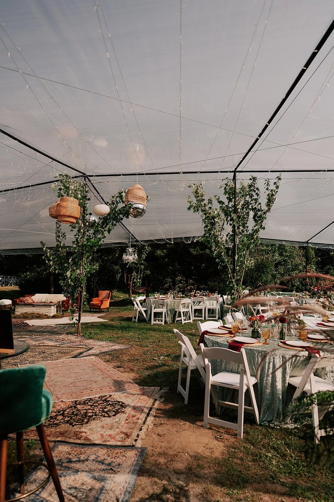 Outdoor party setup with round tables covered in green tablecloths, white chairs, decorative vases, and hanging lanterns under a large canopy decorated with string lights and greenery.