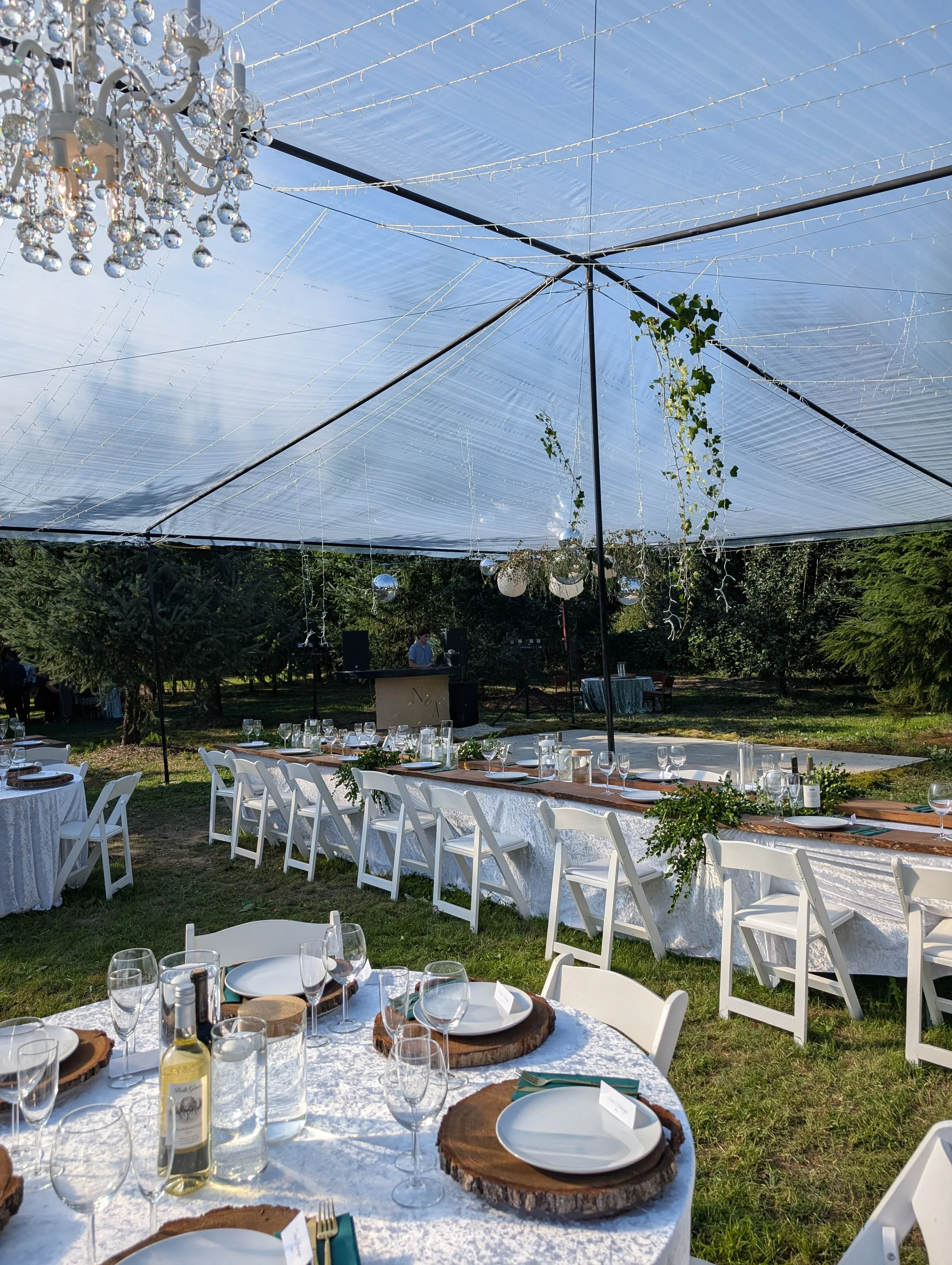 Under a small outdoor tent with string lights, decorated for a wedding or special event, with long tables covered in white tablecloths, set with plates, glasses, and wooden chargers, surrounded by white chairs, and a DJ in the background.
