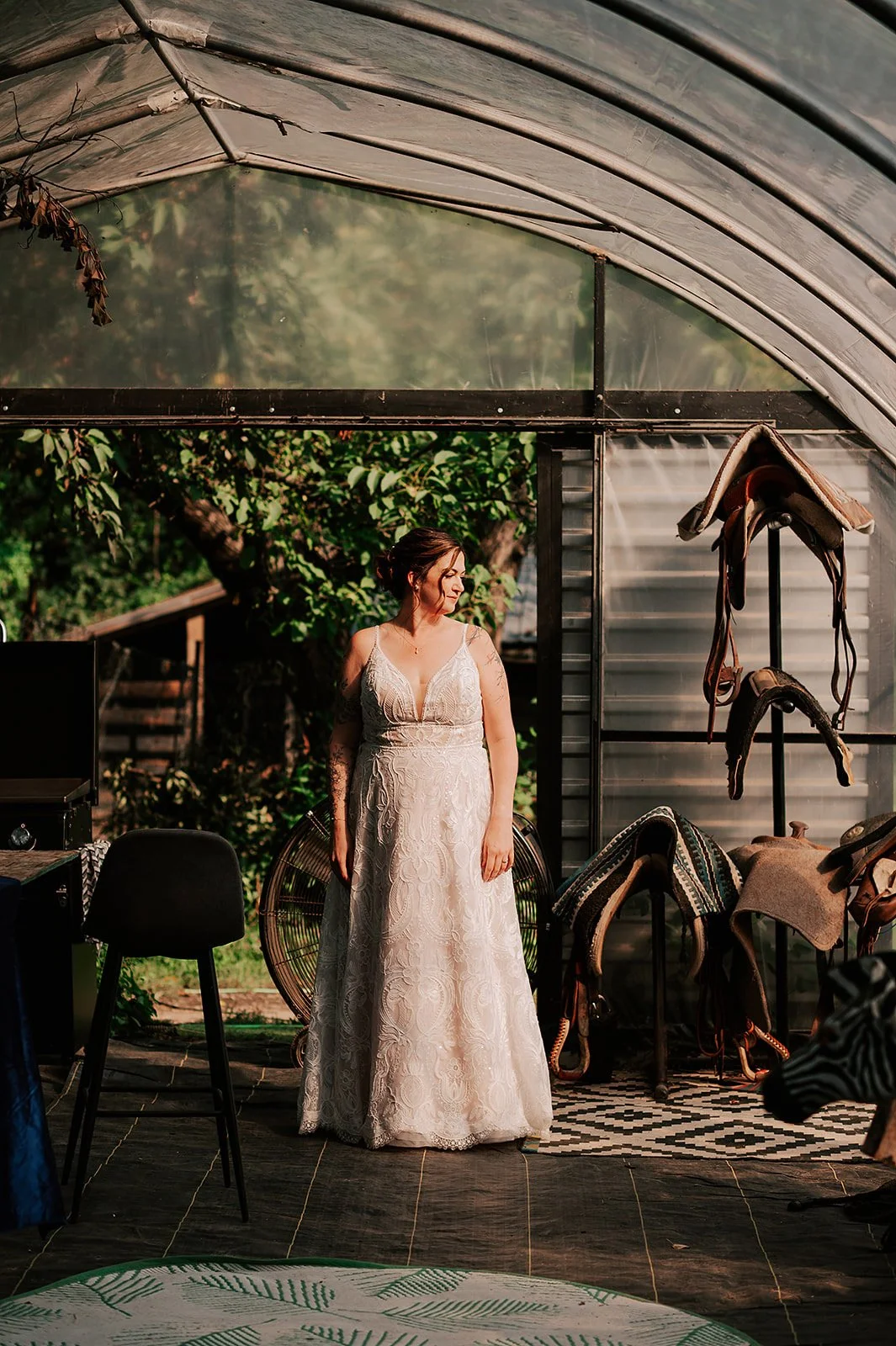 A woman in a white lace wedding dress standing inside a greenhouse or garden structure surrounded by trees and sunlight.