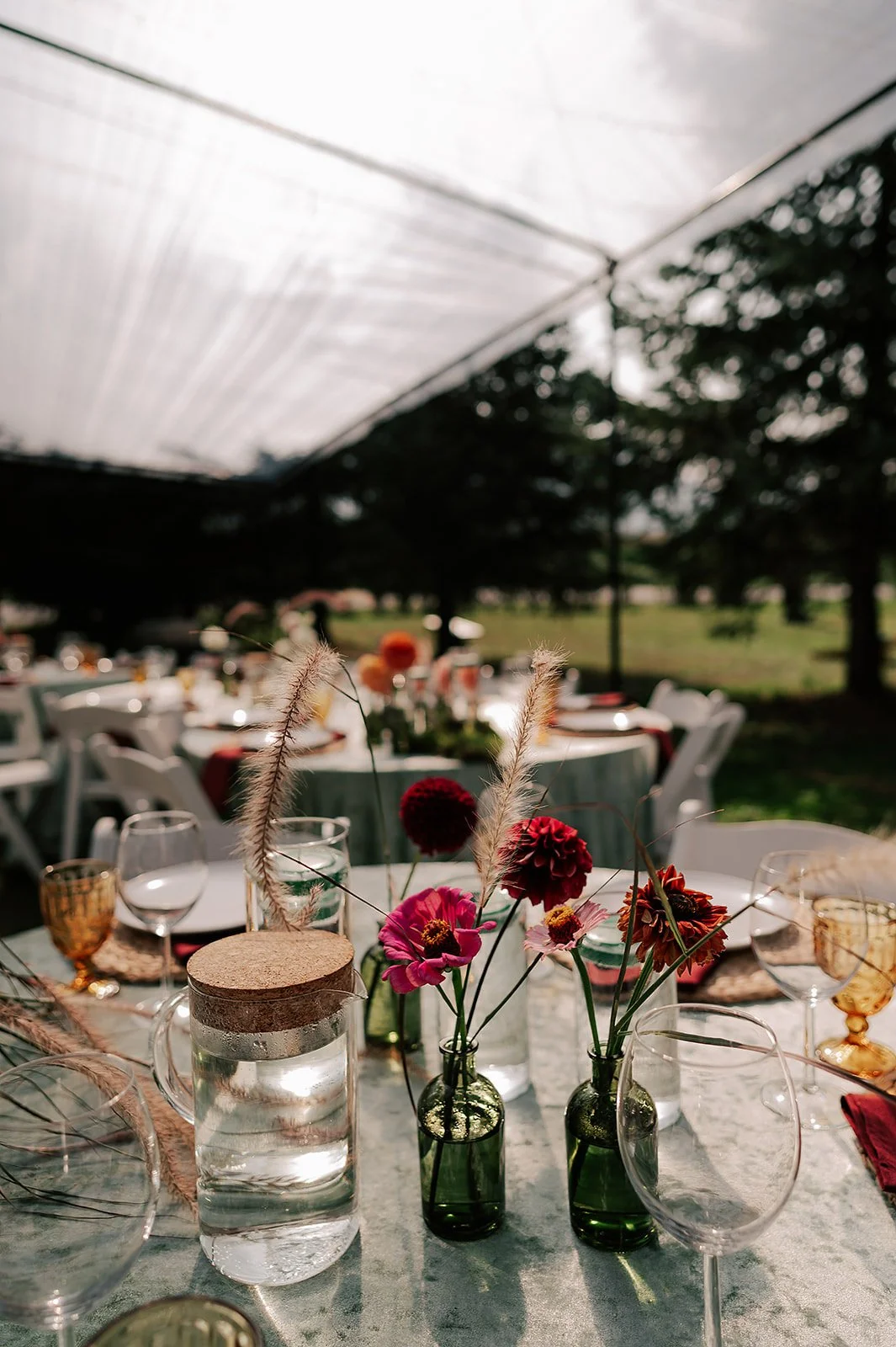 Table set for an outdoor celebration with floral centerpieces in small green glass bottles, water pitchers, wine glasses, and gold-rimmed glasses under a canopy with a background of trees.