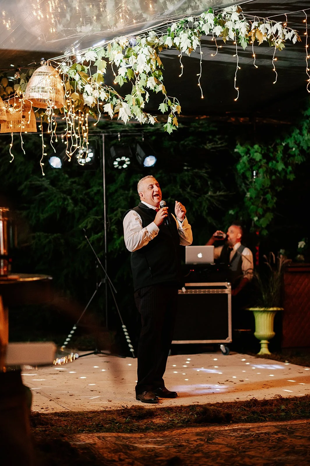 A man singing into a microphone on a decorated outdoor stage at night, with a laptop and DJ equipment behind him, under hanging lights and foliage.
