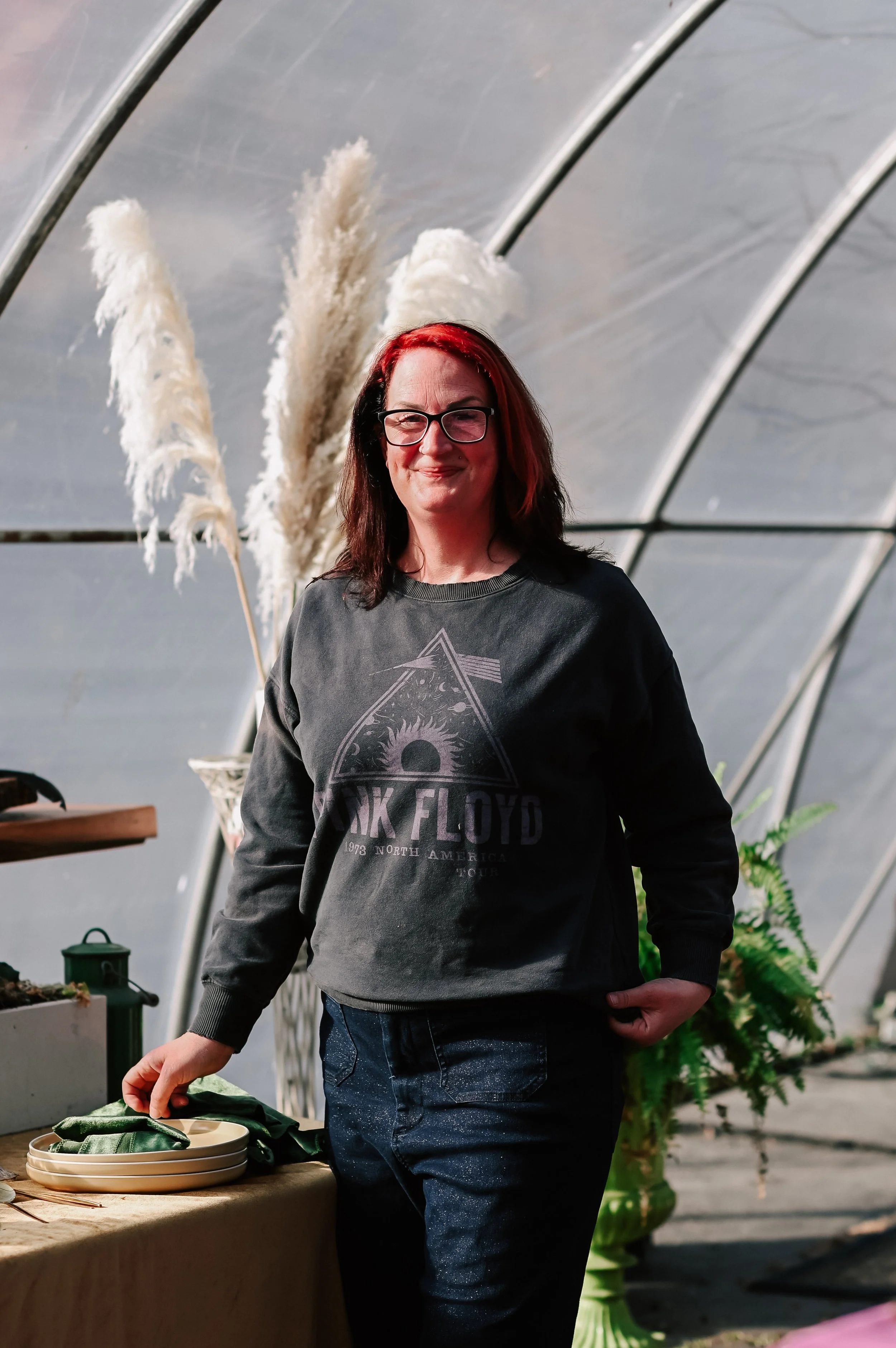 A woman with red hair and glasses standing in a greenhouse, wearing a Black Floyd sweatshirt, with pampas grass and potted plants around her.
