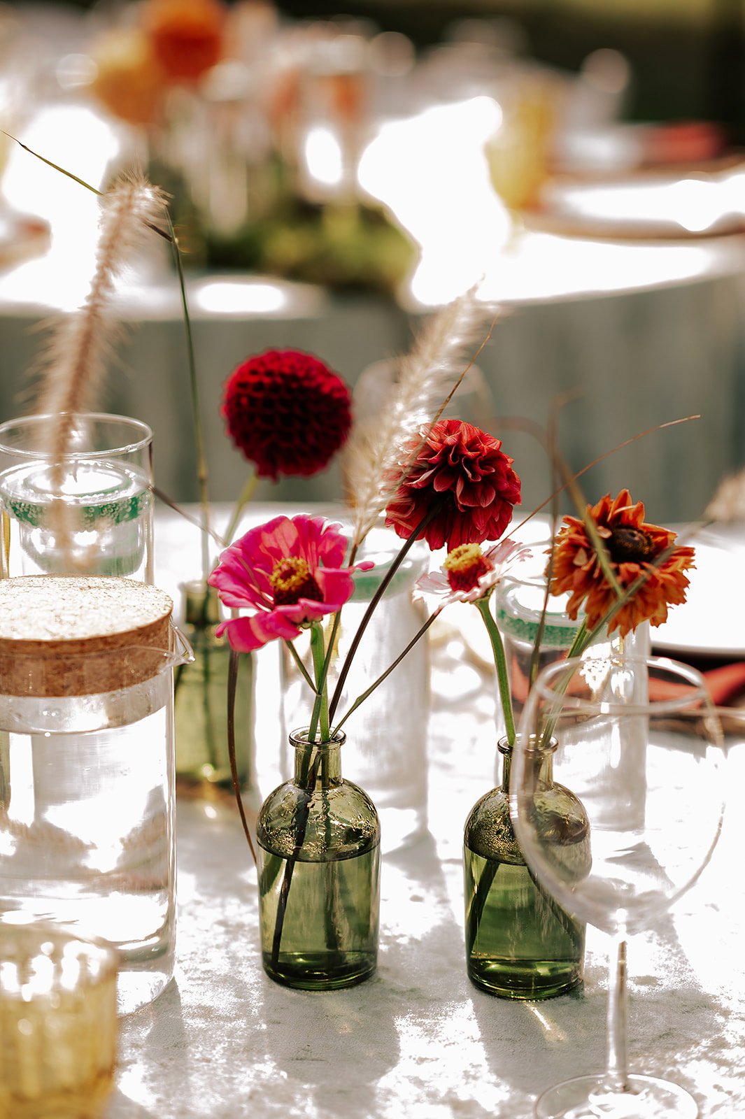 A table with vases holding colorful flowers and decorative grasses, set in bright natural light.