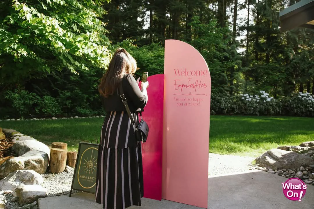 Woman taking a photo with her phone next to a pink and red sign that reads "Welcome" and "Empowerfory" in a garden setting.