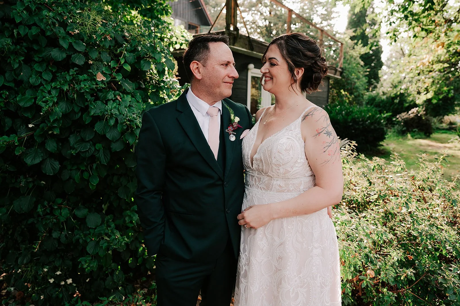 A couple dressed in wedding attire standing close together outdoors amid greenery, smiling at each other.