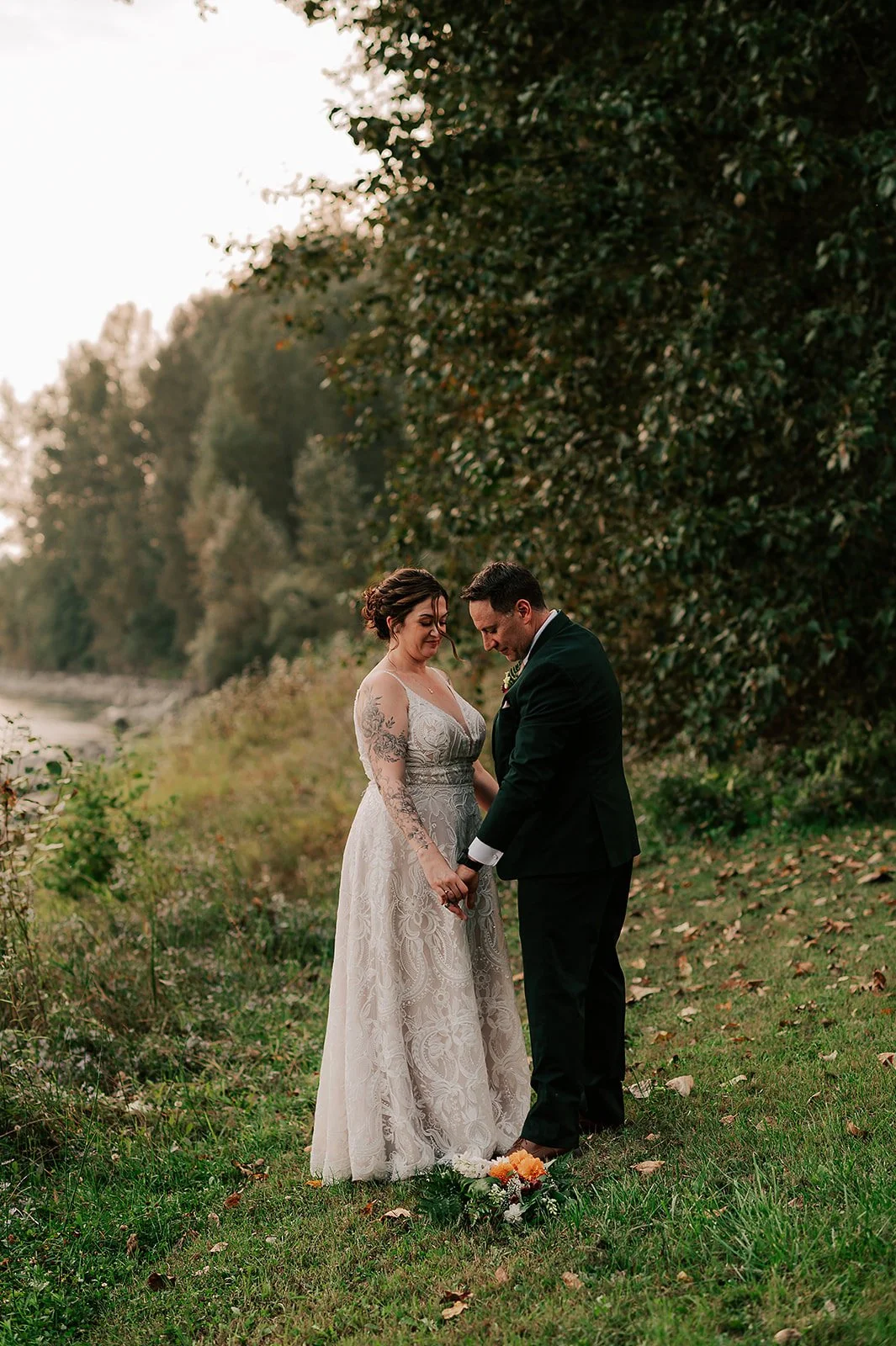 A couple in wedding attire holding hands outdoors by a river, with trees in the background and a bouquet on the grass.