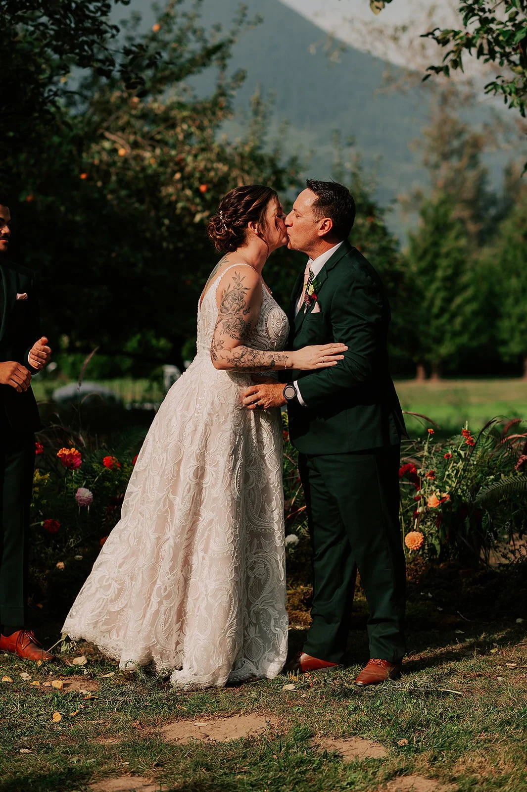 A couple kisses during their outdoor wedding ceremony, with a woman in a white lace dress and a man in a dark green suit, surrounded by greenery and flowers.