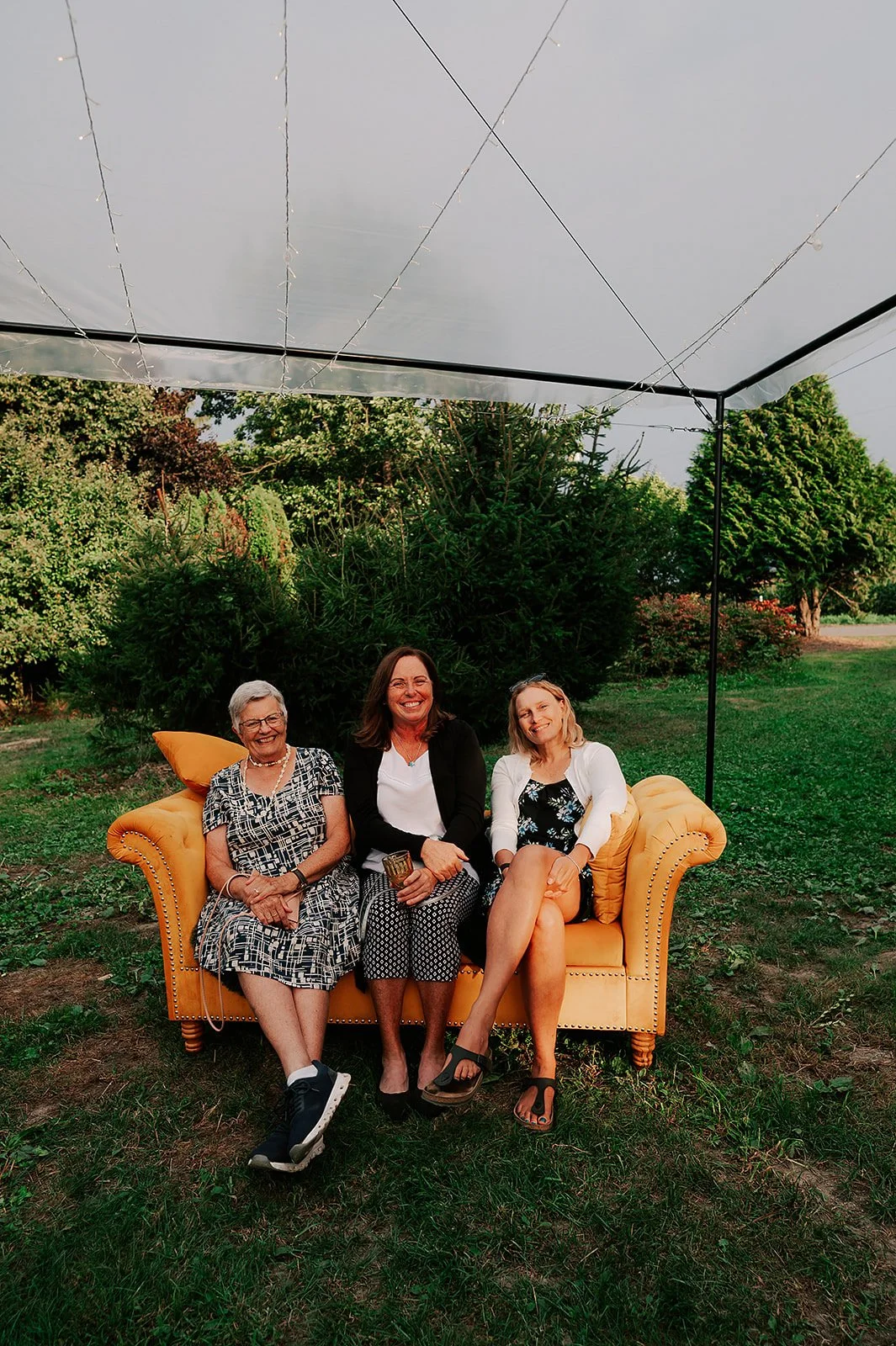 Three women sitting on a yellow sofa outdoors under a small canopy, surrounded by trees and greenery, smiling and enjoying the moment.
