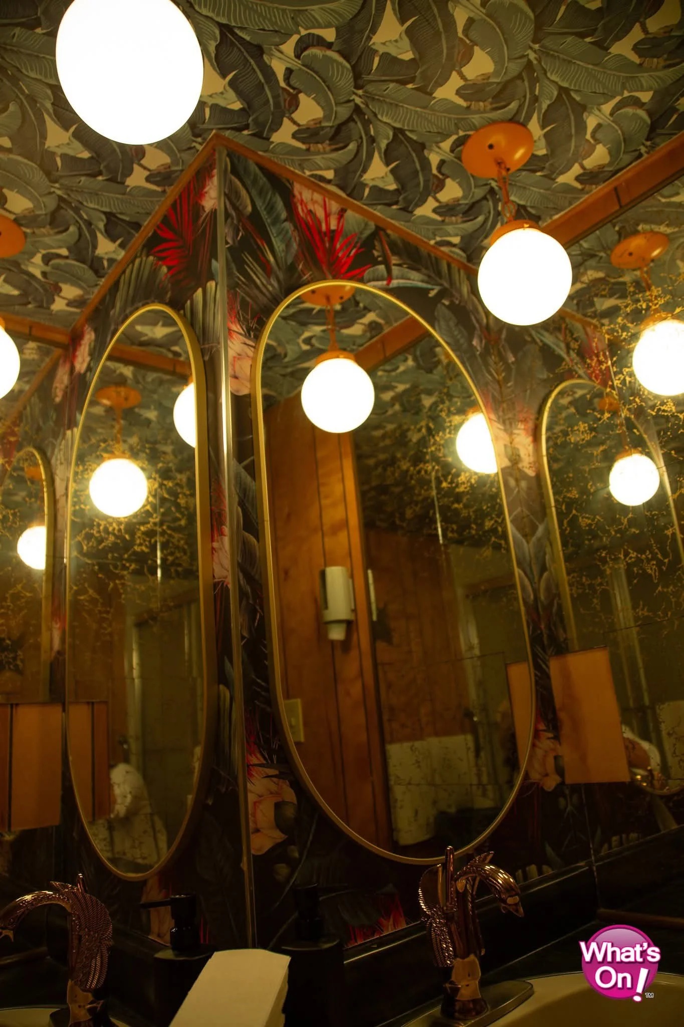 Decorative bathroom mirror with tropical-themed wallpaper, round ceiling lights, wooden walls, and a lamp with a black base, in a cozy interior.