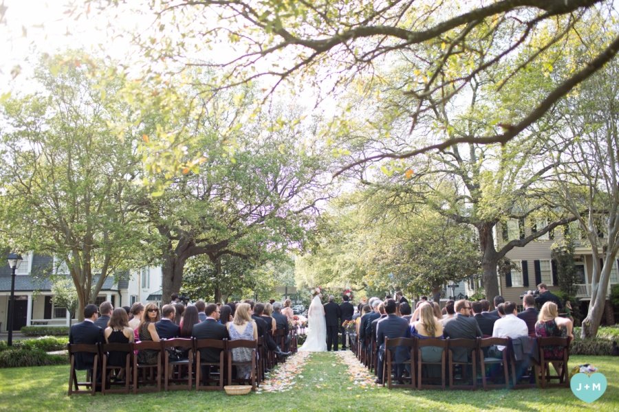 Outdoor wedding ceremony with guests seated on wooden chairs on both sides of a grassy aisle, under green trees in a residential neighborhood.