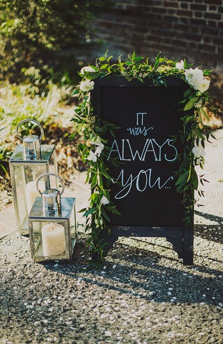 A chalkboard sign with a greenery and white flower border, reading 'It was always you...' set outdoors on a gravel surface. Two lanterns with candles are placed beside it.