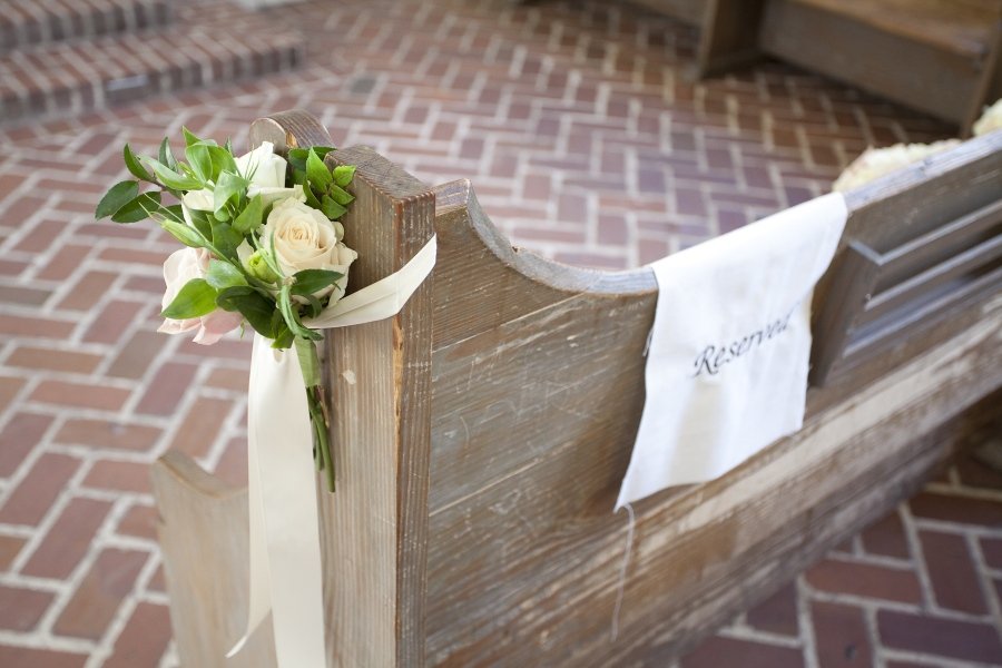 A bouquet of white and pale pink roses with green leaves attached to the end of a wooden church pew, which has a white cloth with 'Reserved' embroidered on it. The background shows a brick floor.
