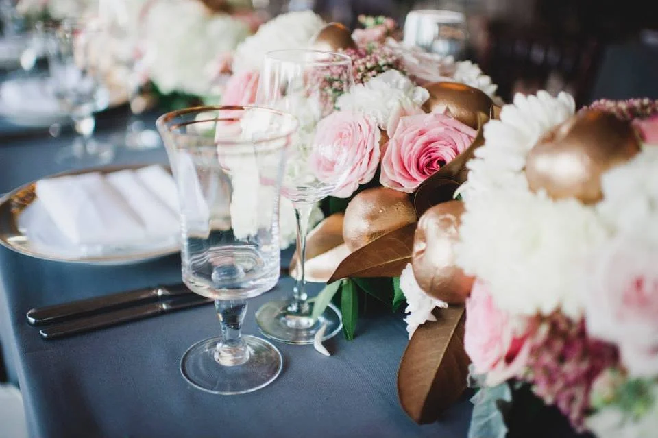 Table setting with clear wine glasses, a floral centerpiece featuring pink roses and white flowers, and gold accents, suggesting a formal event or celebration.