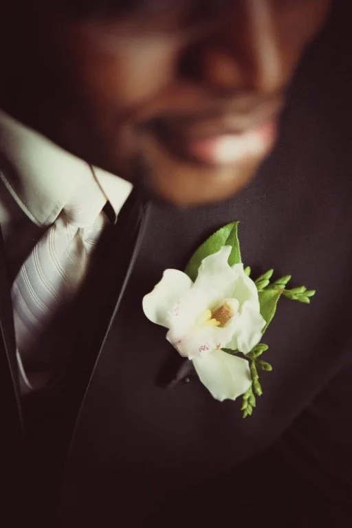 Close-up of a man in formal attire with a white flower boutonniere on his dark jacket, smiling softly.