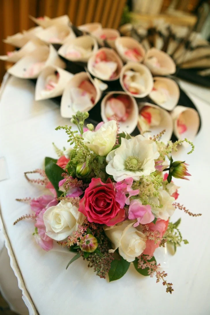 Close-up of a floral arrangement with white, pink, and red roses, and other small flowers, in front of a table with numerous rolled-up paper cones filled with flower petals.