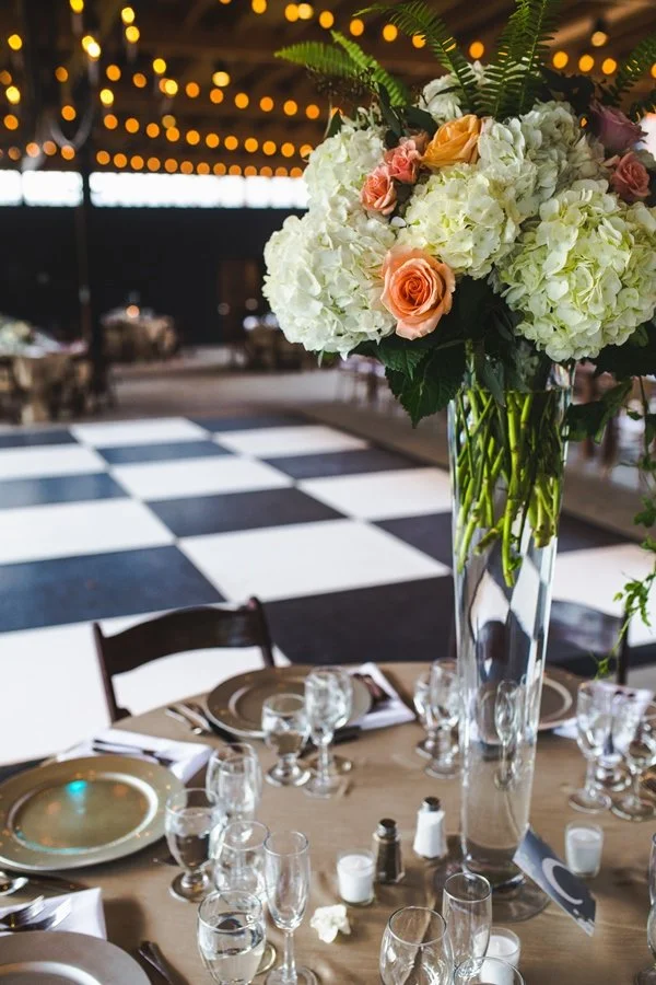 A dining table setup with a large floral centerpiece in a tall glass vase, dressed with white hydrangeas and peach roses, in a decorated venue with string lights and a checkered dance floor in the background.