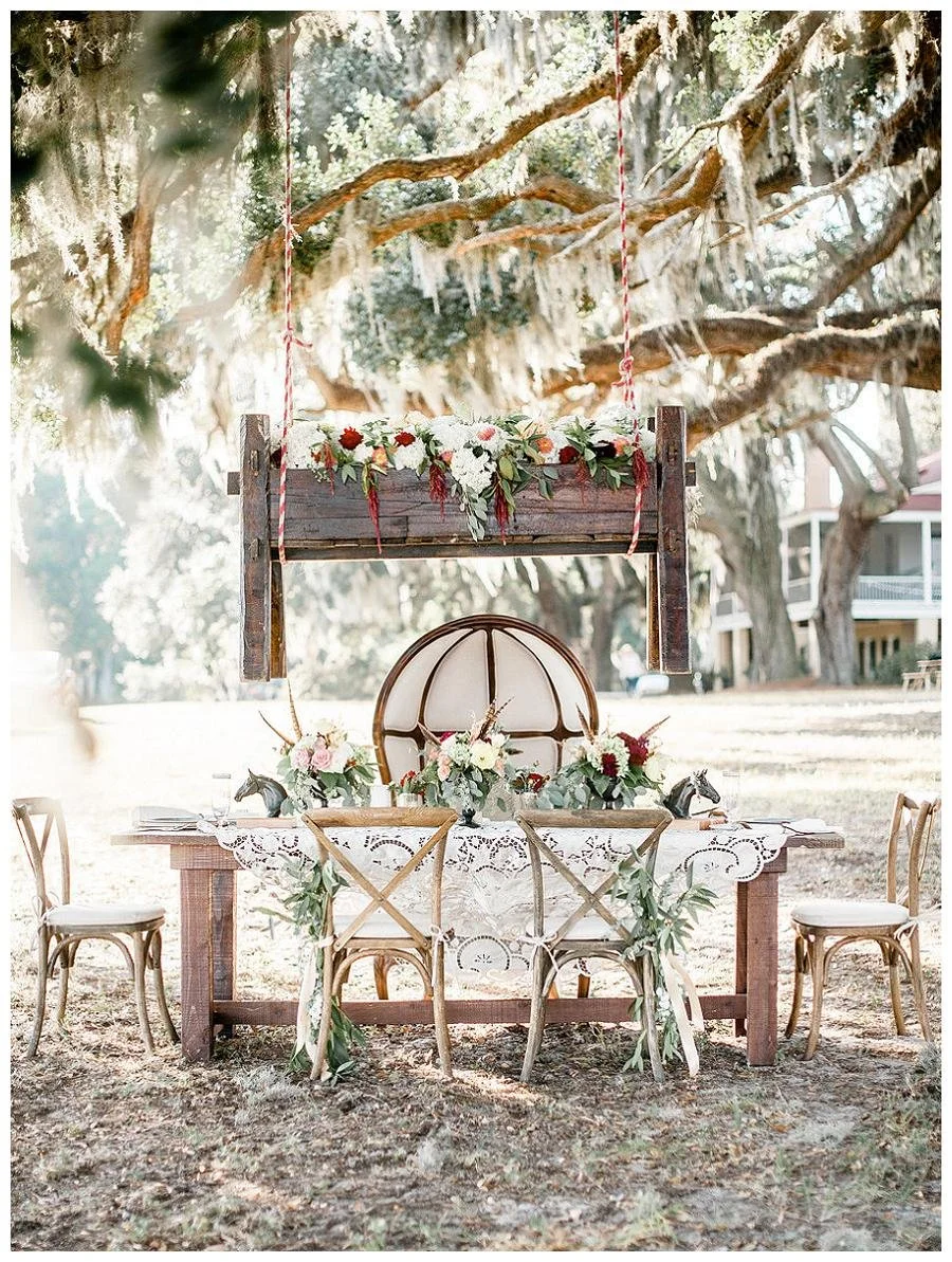 Outdoor table decorated for a rustic wedding with floral arrangements, surrounded by chairs, under a large tree with hanging moss.