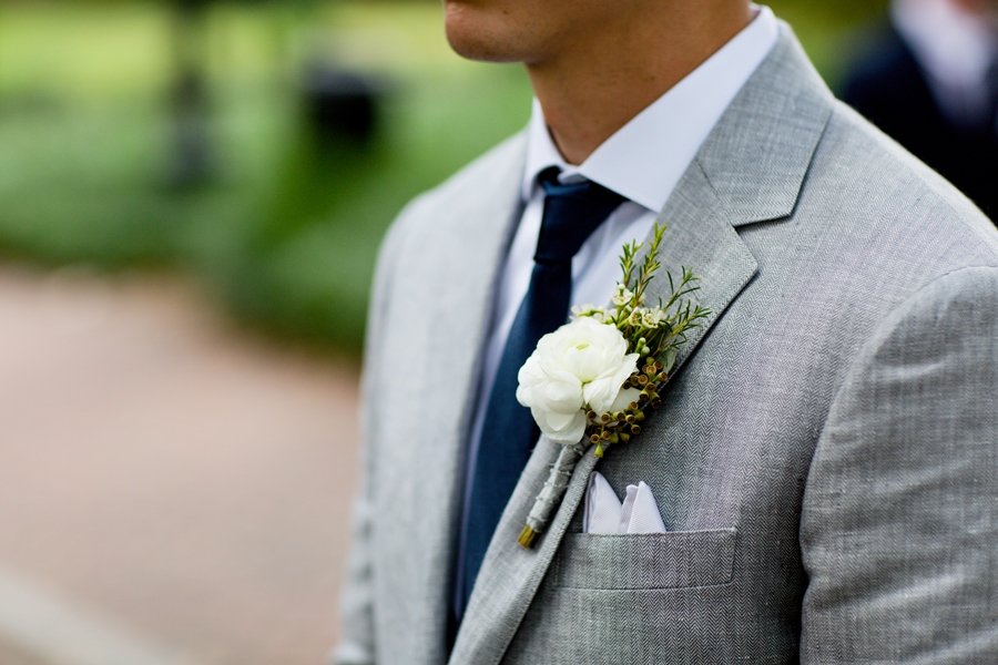 Close-up of a man wearing a light gray suit with a white shirt and navy tie, showcasing a boutonniere of white flowers and greenery on his lapel, likely at a formal event outdoors.