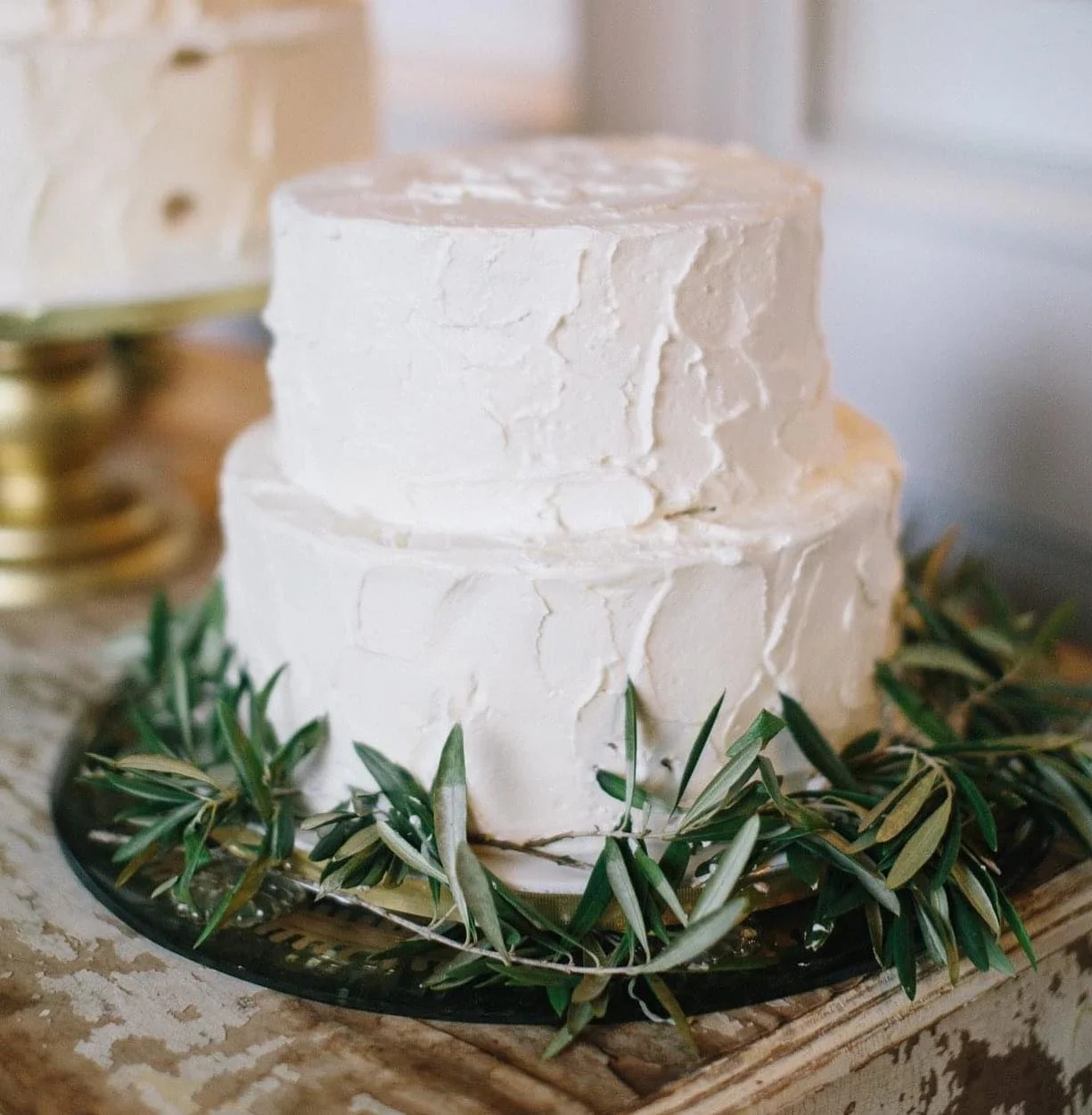 Two-tiered white frosted cake on a black platter decorated with green olive branches.