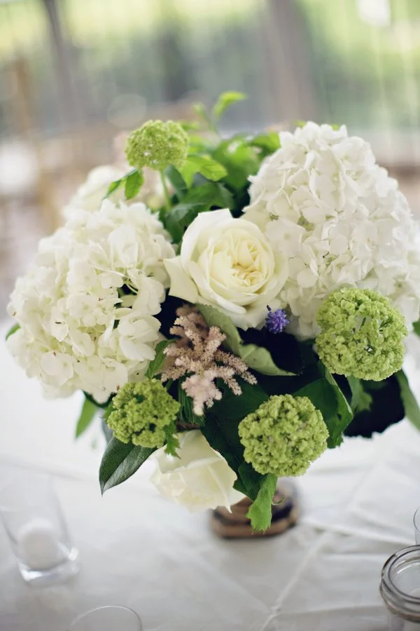 A floral arrangement with white roses, white hydrangeas, green ball-shaped flowers, and small purple accents, set in a vase on a table.