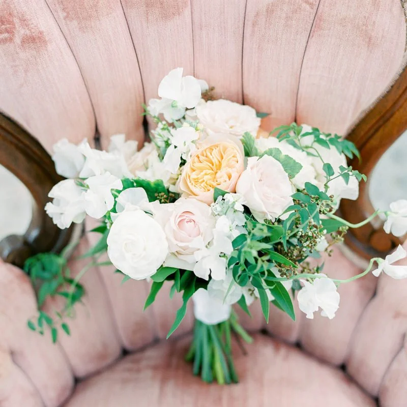 A bouquet of assorted flowers including white, blush, and peach roses, placed on a pink upholstered chair with wooden accents.