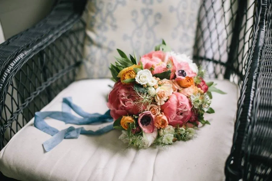 A bouquet of mixed flowers placed on a cushioned bench between black wicker chairs, with a blue ribbon tied to it.