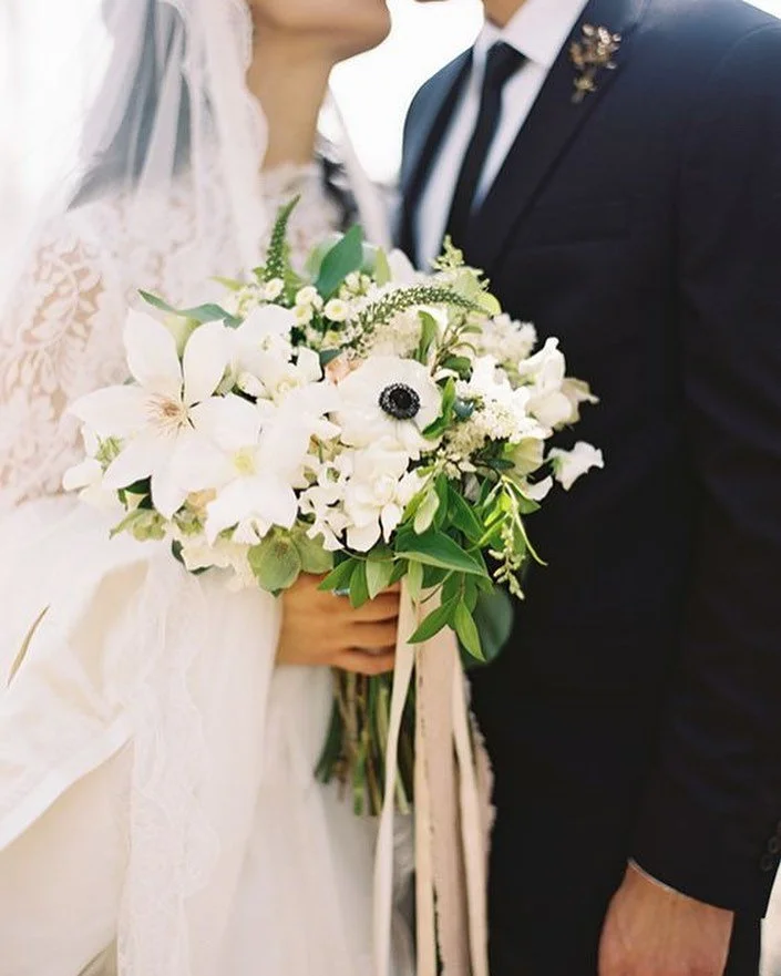 Bride holding a bouquet of white flowers standing next to groom in a dark suit, wedding setting.