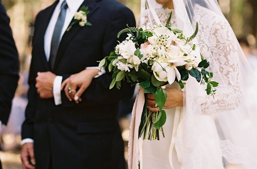 A bride in white lace holding a bouquet of white flowers and greenery at a wedding ceremony.