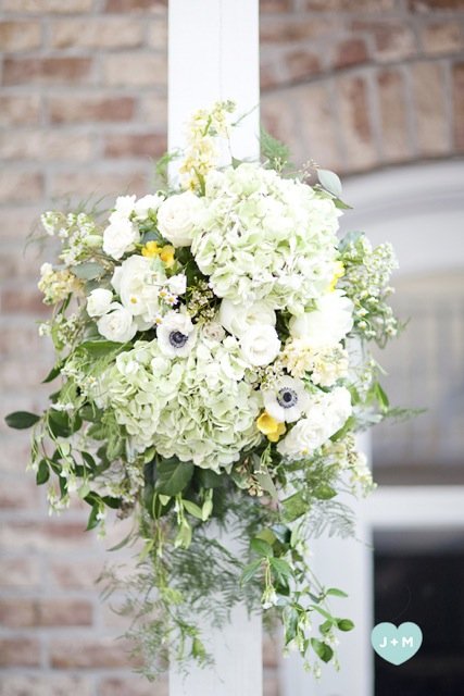 White floral arrangement with hydrangeas, roses, and greenery hanging from a white post.