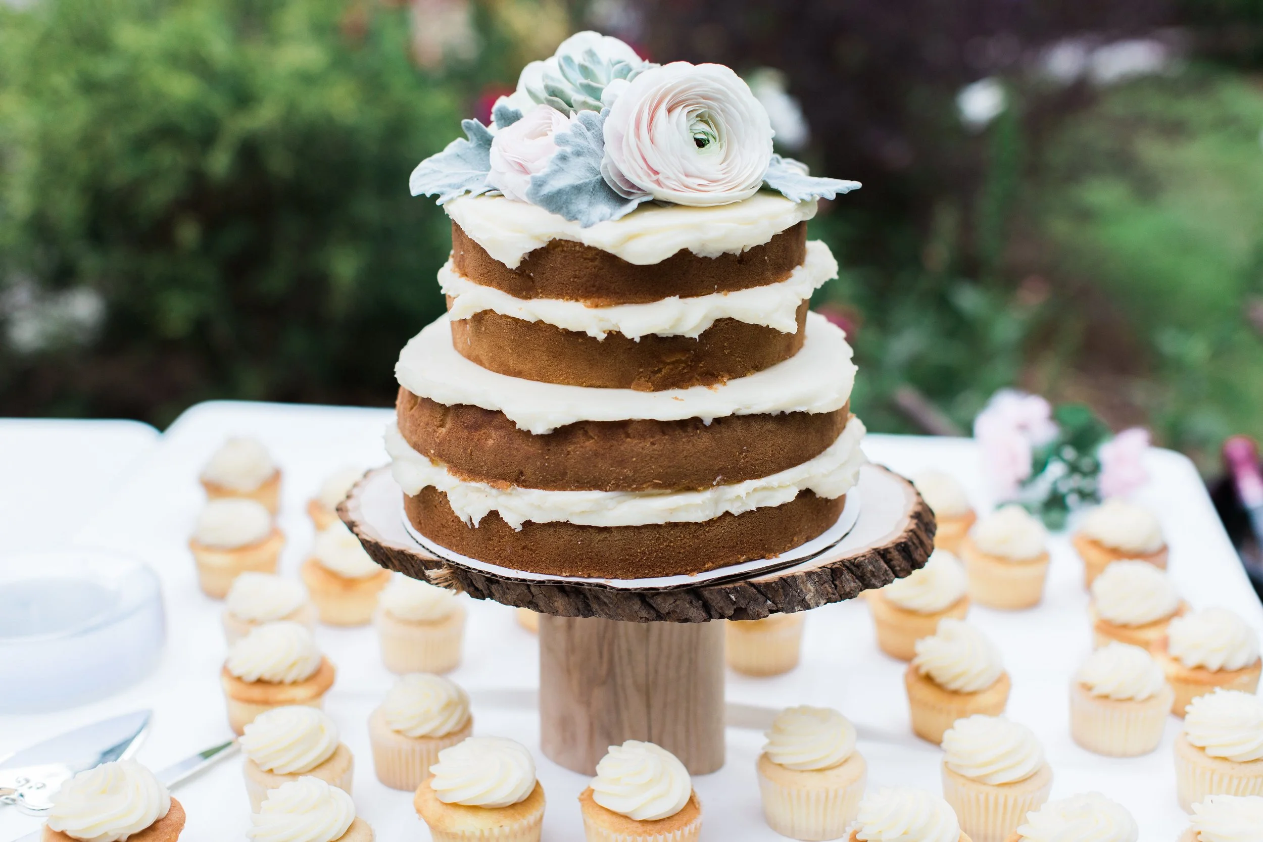 A multi-layered semi-naked cake with white icing and flowers on top, surrounded by cupcakes with white frosting, set on a rustic cake stand outdoors.