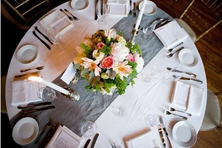 Round table set for a formal dinner with a floral centerpiece, candles, white plates, silverware, and wine glasses, with a gray table runner.