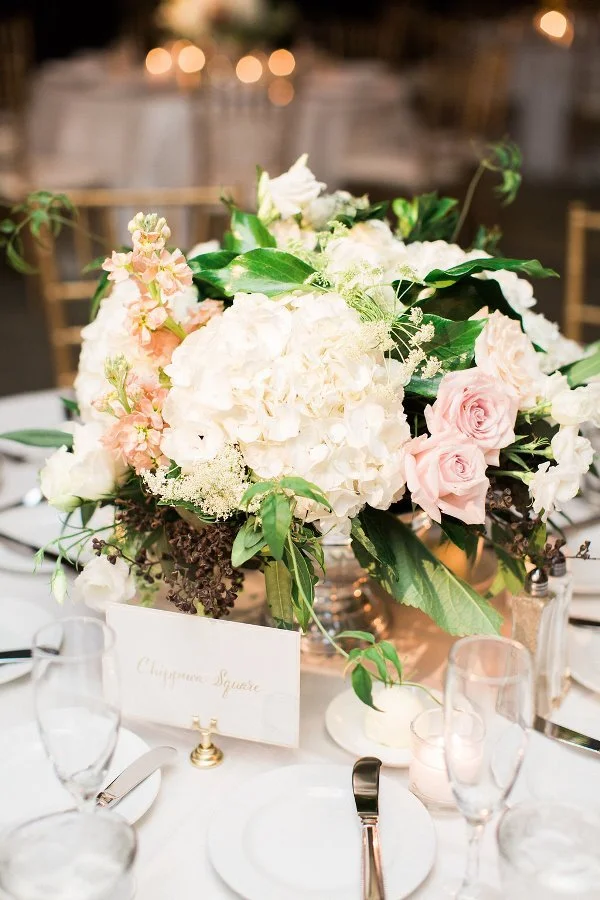 Elegant wedding table centerpiece with white and pale pink flowers and lush greenery, set on a white table with glassware and a table card.