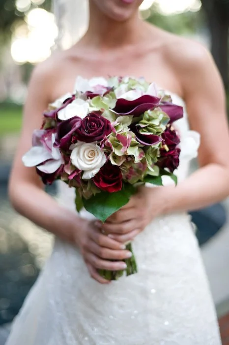 Bride holding a bouquet of white, deep red, and light green flowers outdoors