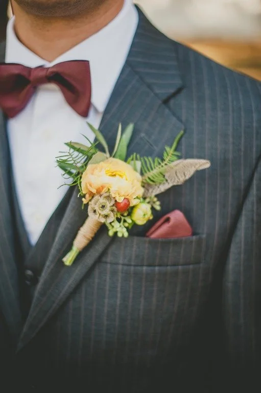 Close-up of a man wearing a dark pinstripe suit, white shirt, maroon bow tie, and pocket square, with a boutonniere pinned to his lapel featuring a peach-colored flower, green foliage, and small decorative elements.