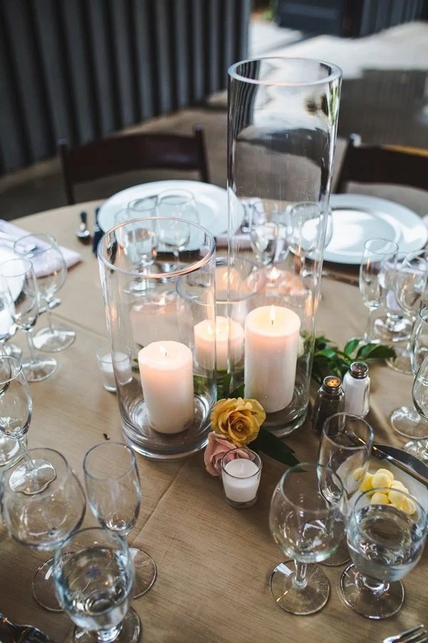 Decorative table setting with three candles inside glass holders, a yellow rose, and a small candle on a beige tablecloth, with plates, silverware, and glasses arranged around.