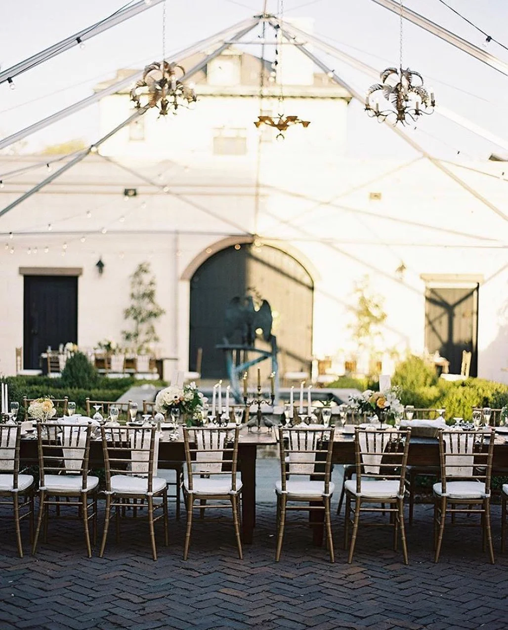 Elegant outdoor dining setup with a long wooden table adorned with floral centerpieces, surrounded by wooden chairs with white cushions under a transparent tent with chandeliers and string lights. In the background, a white building with black doors 