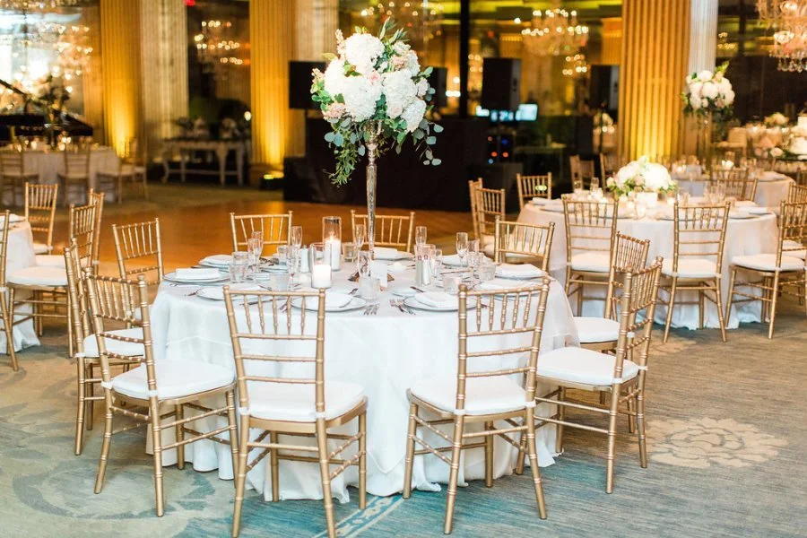 Elegant banquet table with white tablecloth, gold chairs, and floral centerpiece in a decorated event hall with warm lighting.