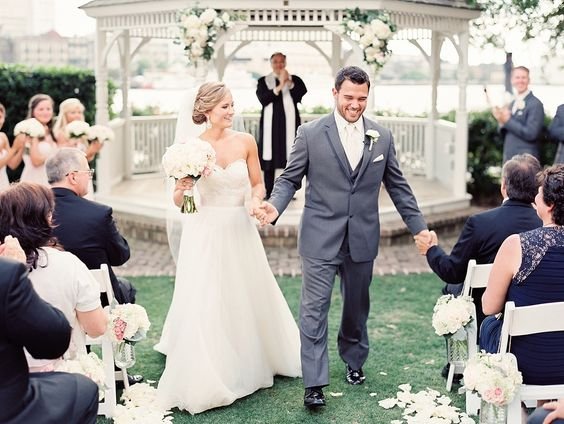 A couple is having a wedding ceremony outdoors in a white gazebo, with the bride in a white wedding dress holding a bouquet and the groom in a gray suit holding her hand. Guests are seated on white chairs, and there are flower arrangements along the 