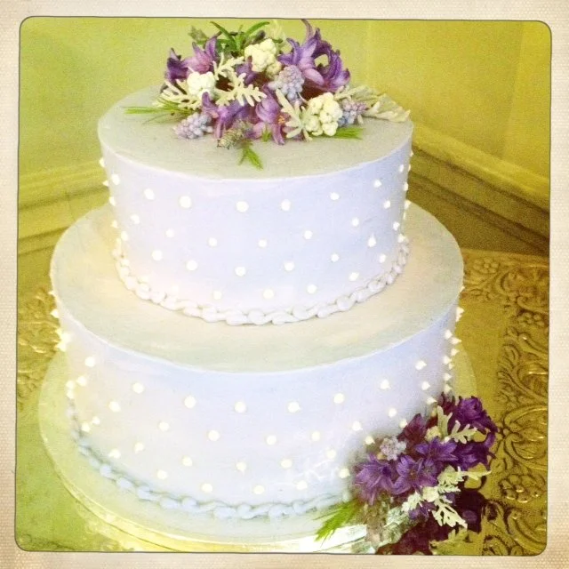 Two-tier white wedding cake decorated with purple and white flowers on top and at the base, with a dotted pattern of white icing