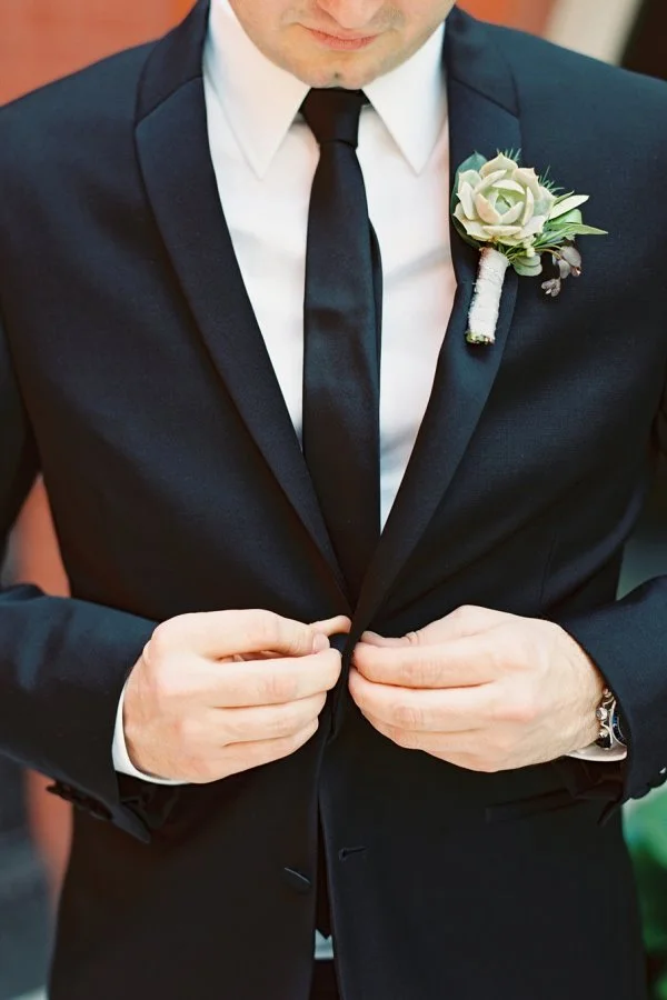Close-up of a groom adjusting his black suit jacket, with a white shirt, black tie, and a white boutonniere on his lapel.