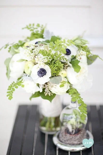A bouquet of white anemones with dark centers, green leaves, and small white flowers in a white vase on a dark wooden table.